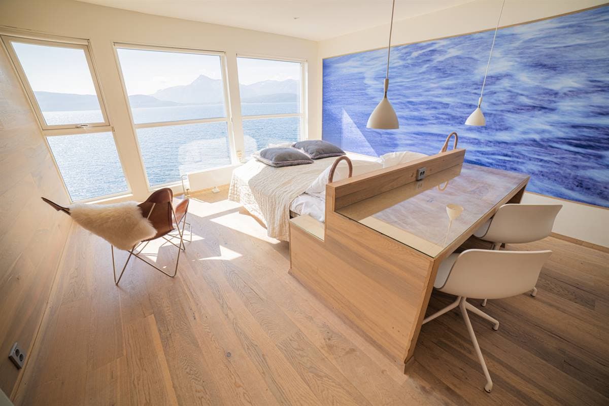 Skrivebord og seng under et vægmaleri med bølger samt vinduer ud mod fjorden
