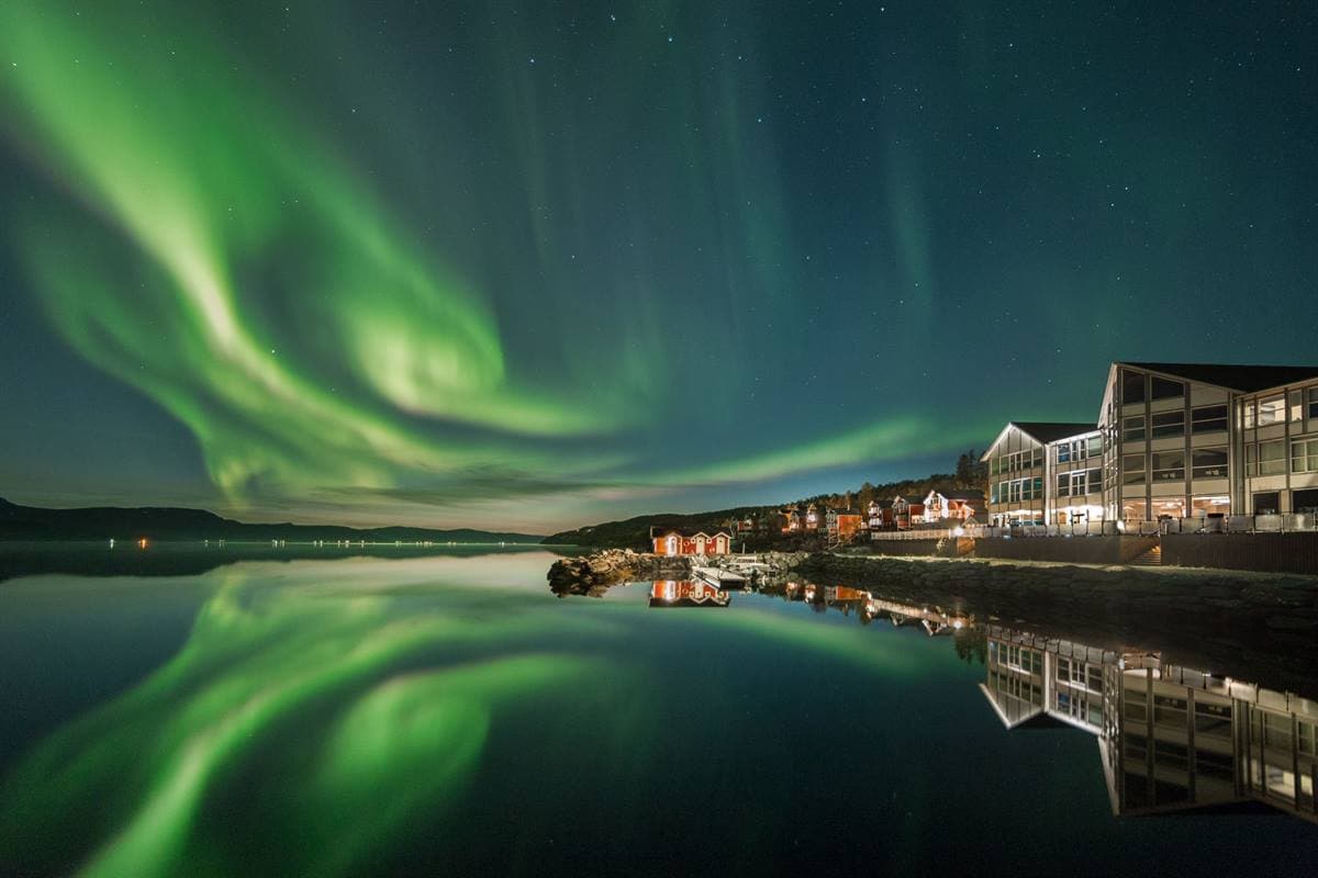 Nordlys over Malangenfjorden, der spejler sig i vandet ved det oplyste resort